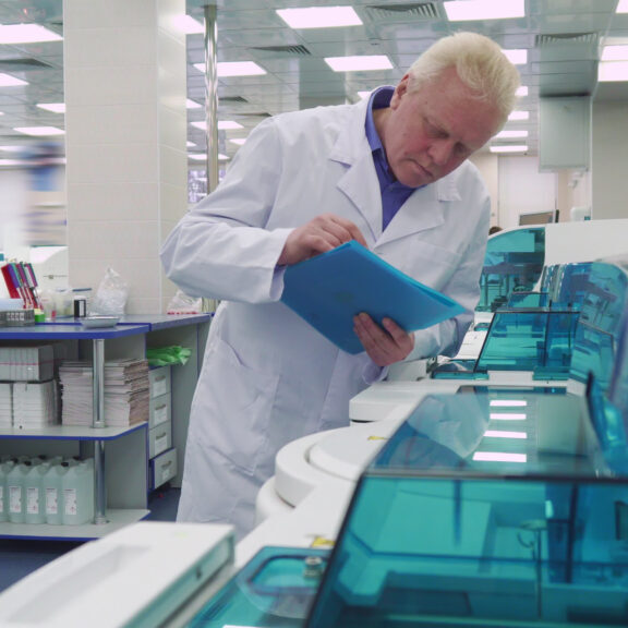 Gray aged man standing near laboratory devices. Senior lab worker looking at scientific equipment. Mature male scientist standing on background of working centrifugal machine