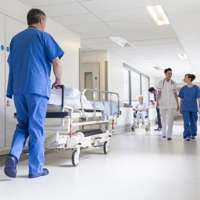 Male nurse pushing stretcher gurney bed in hospital corridor with doctors &amp; senior female patient