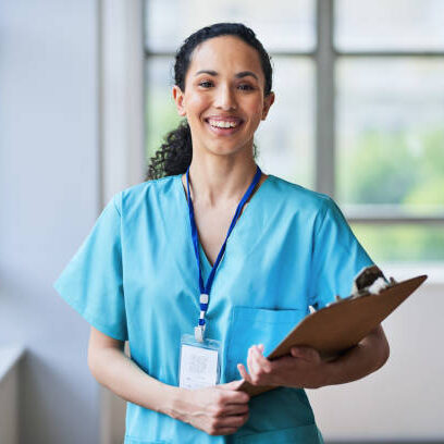 Portrait of a cheerful medical professional in blue scrubs holding a clipboard. The multi-ethnic healthcare worker is standing in a bright hospital hallway, symbolizing diversity, dedication, and expertise in the medical field.